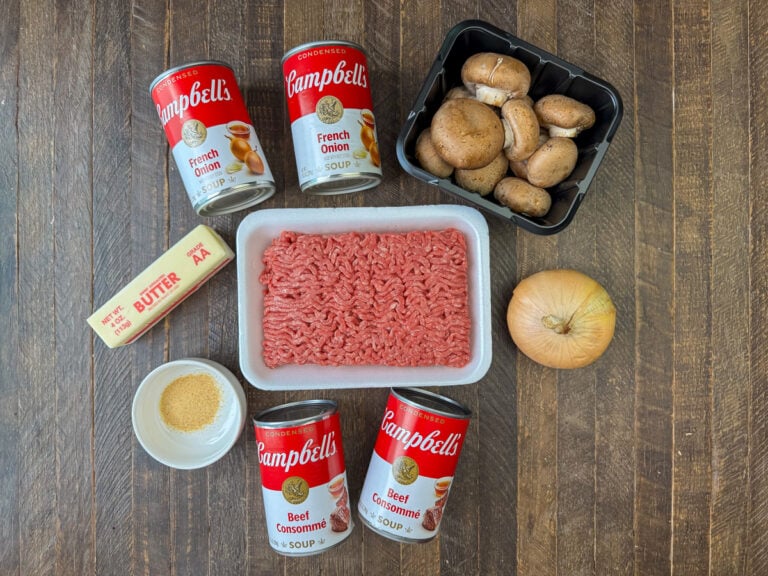 Ingredients to make a beef and rice casserole are cans of French onion soup, baby bella mushrooms, ground beef, an onion, a stick of butter, garlic powder and cans of beef consomme on a wooden counter top.