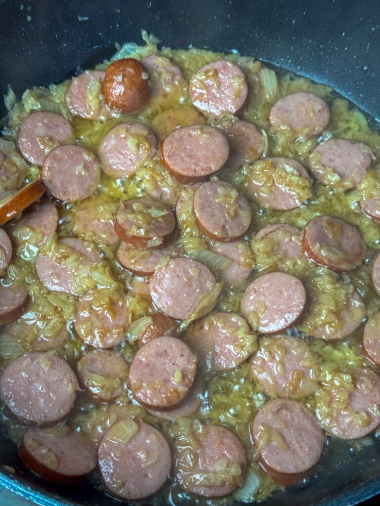 Coins of Kielbasa sausage cooking in a skillet with chopped onions saut&eacute;ing in butter.