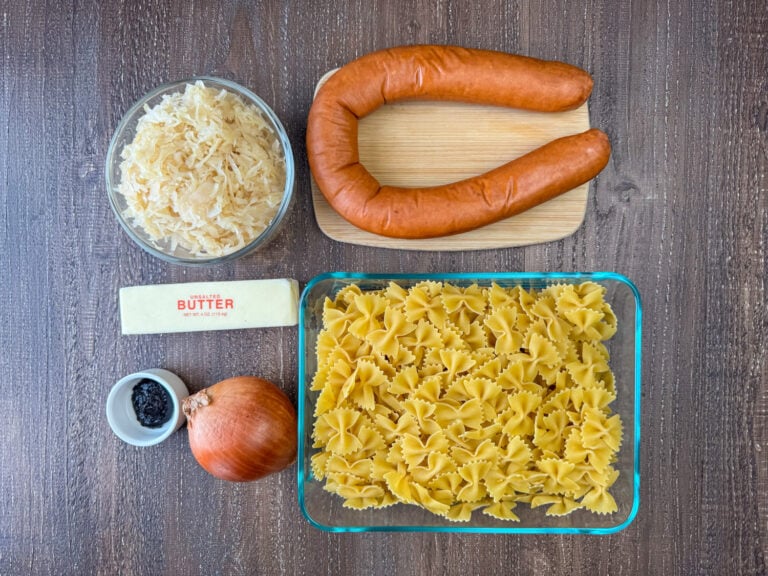 Ingredients to make Kielbasa and pasta are butter, sauerkraut, Kielbasa sausage, bow tie pasta, onion and beef bouillon paste on a wooden counter top.