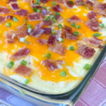 Close up casserole dish with loaded mashed potatoes topped with bacon, two types of cheese and green onion slices.