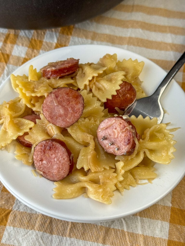 Fork in pieces of bowtie pasta and a slice of Kielbasa accented with sauerkraut and onion ready to take a bite on a plate full of the easy meal.