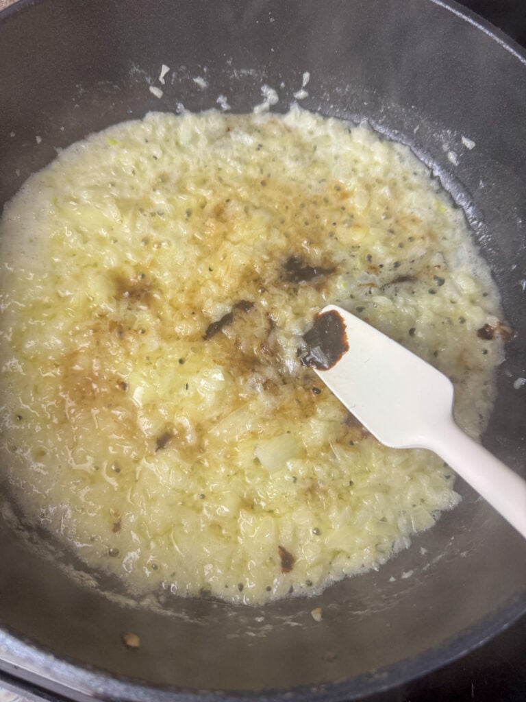 Spatula stirring beef broth paste and mustard into a skillet of onions softened in butter.