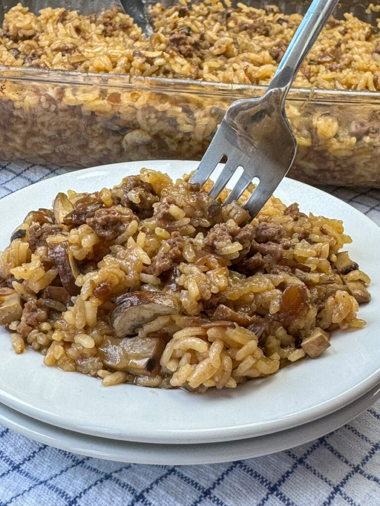 Butter, Consomme and French onion soup coating every grain of rice with ground beef and a few mushrooms nestled in a scoop of this casserole on a white plate with the dish in the background.