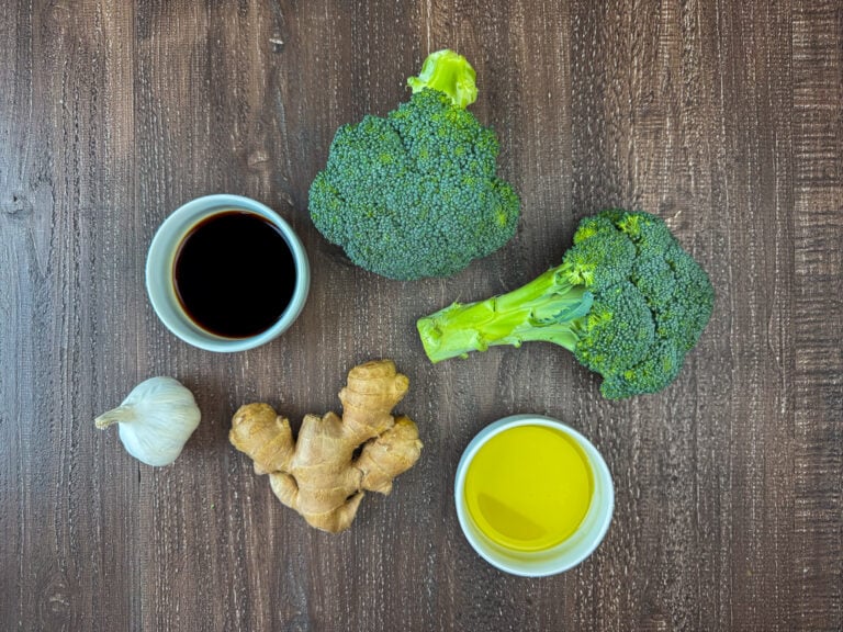 Ingredients to make Asian broccoli are two bunches of broccoli, olive oil, ginger, garlic and soy sauce on a wooden counter top.