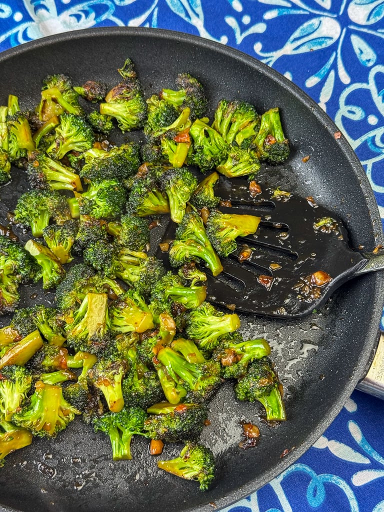 Spatula scooping up tender, stir fried Asian broccoli with soy sauce out of a skillet on a bright blue and white napkin.