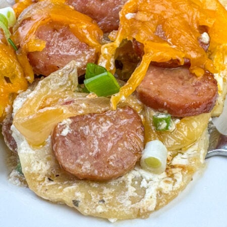 Close up cheesy Pierogi enhanced with Kielbasa, onions and sour cream while baking in beef broth with a fork underneath.