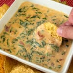 Close up of a hand dipping a chip in a delicious, warm, creamy spinach dip with bacon.