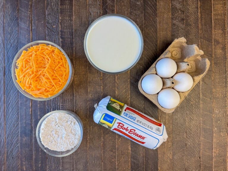 Ingredients to make a breakfast souffl&eacute; are shredded cheddar cheese, milk, eggs, sausage and baking mix on a wooden counter top.