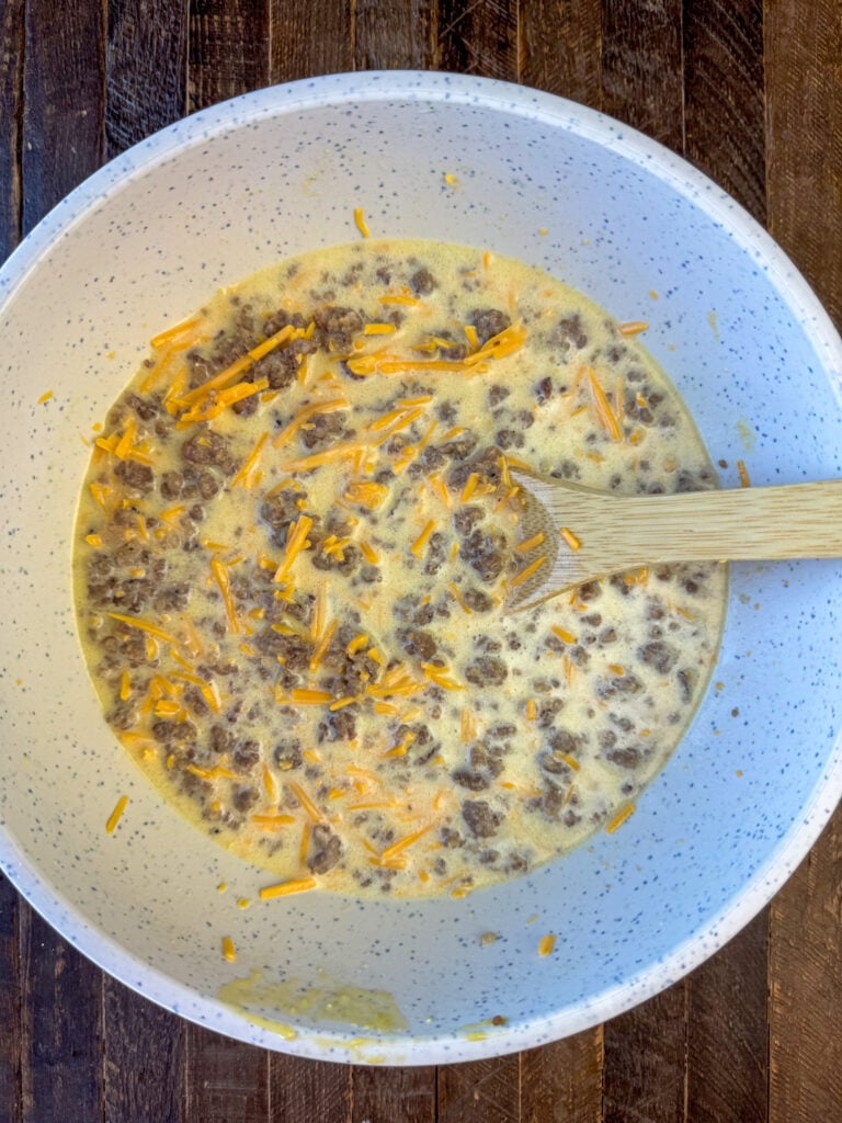 Wooden spoon mixing crumbled sausage and cheddar cheese into an egg and Bisquick mixture in a gray bowl.