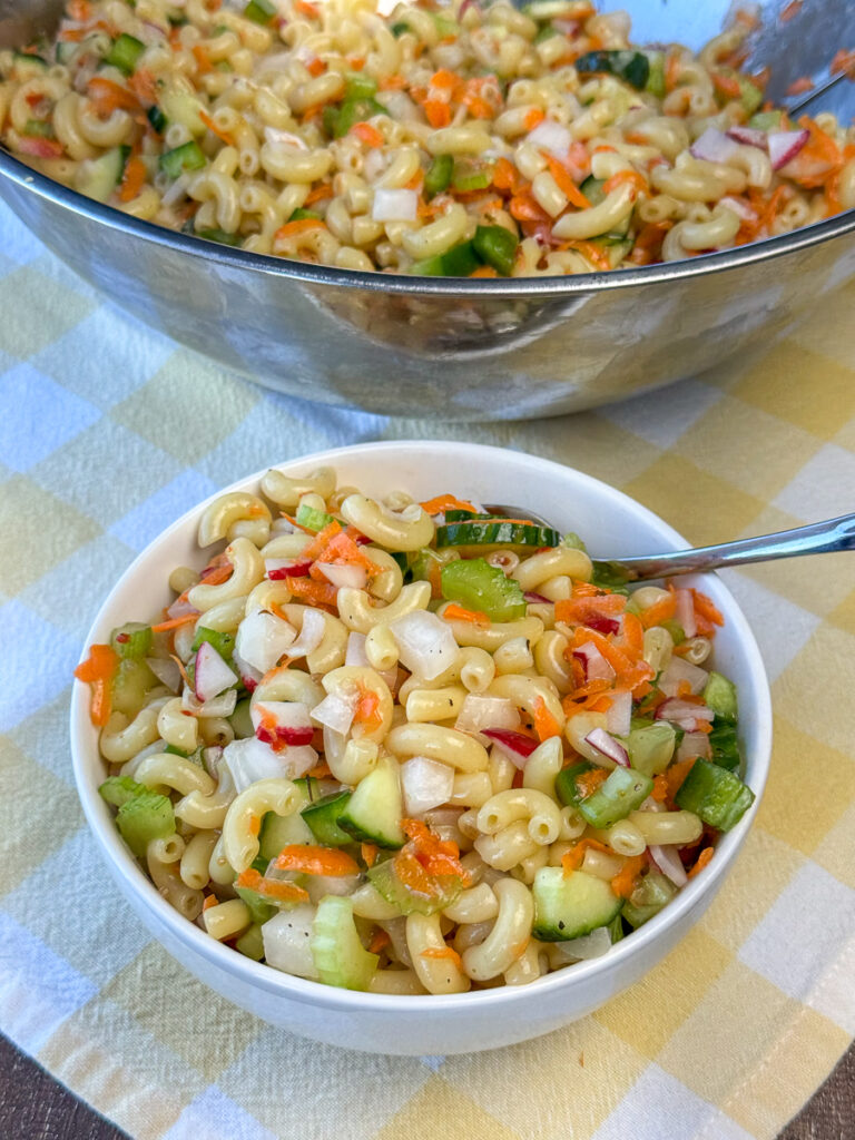 Small white bowl of sweet and flavorful, old fashioned macaroni salad on a yellow and white plate napkin in front of the full serving bowl full.
