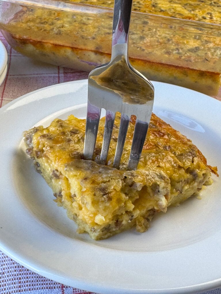 Fork in a piece of delicious egg sausage souffle ready to take a bite with the full breakfast casserole in the background.