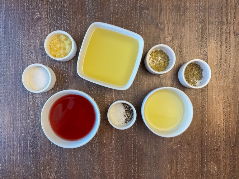 Ingredients to make Greek Dressing are virgin olive oil, red wine vinegar, fresh squeezed lemon juice, garlic, sugar, dried oregano, dried basil, sea salt and freshly ground black pepper on a wooden counter top.