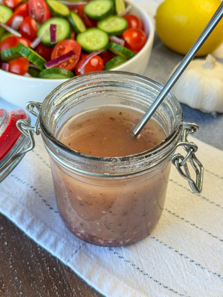 Long handled spoon stirring together a light and enjoyable Greek salad dressing in a glass jar with cucumbers and tomatoes in a bowl in the background and a lemon and bulb of garlic on a blue and white napkin.