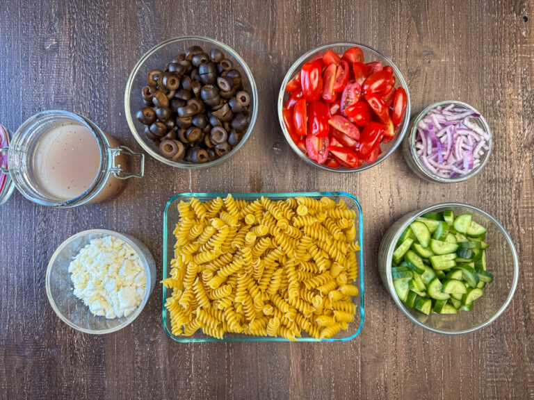Ingredients to make Greek pasta salad are black olives, cherry tomatoes, red onion, cucumber, rotini pasta, feta cheese and Greek salad dressing on a wooden counter top.