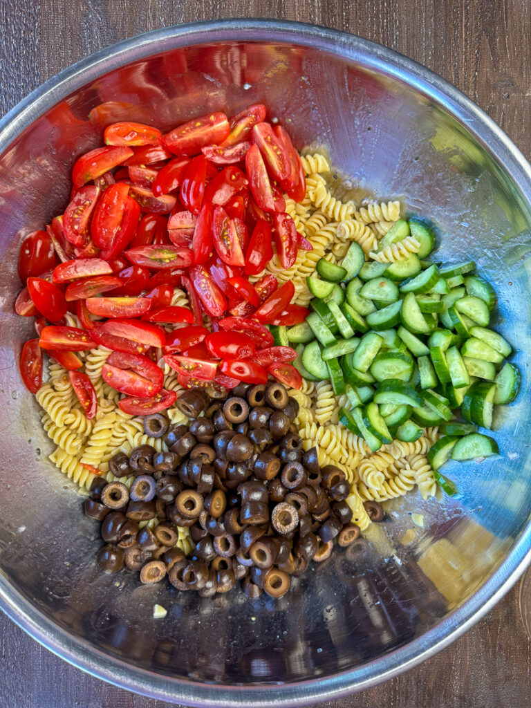 Overhead view of a large mixing bowl with a pound of twist pasta, cherry tomato halves, mini cucumber slices and sliced black olives in sections on top of the pasta.