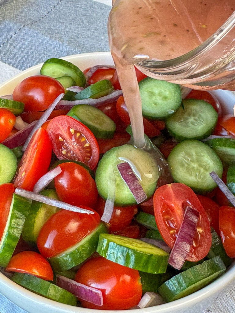 Mason jar with fresh Greek salad dressing pouring the light, fresh recipe over top of sliced cucumbers, tomatoes and red onions in a white bowl.