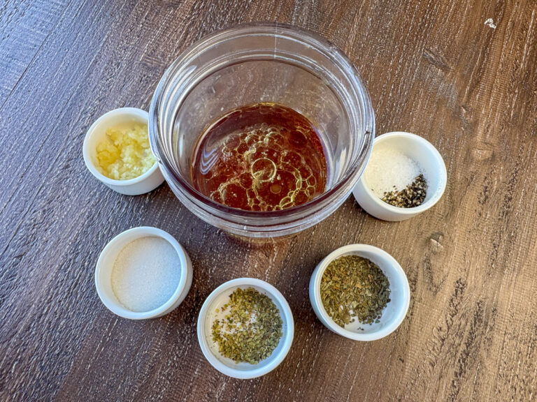 Jar with virgin olive oil and red wine vinegar with small ramekins with minced garlic, sea salt, basil, oregano and sugar on a wooden counter top.