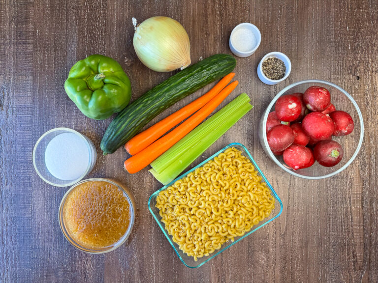 Ingredients to make Italian Macaroni Salad are green pepper or mango, onion, English cucumber, carrots, celery, salt, pepper, radishes, prepared Italian dressing and sugar on a wooden counter.