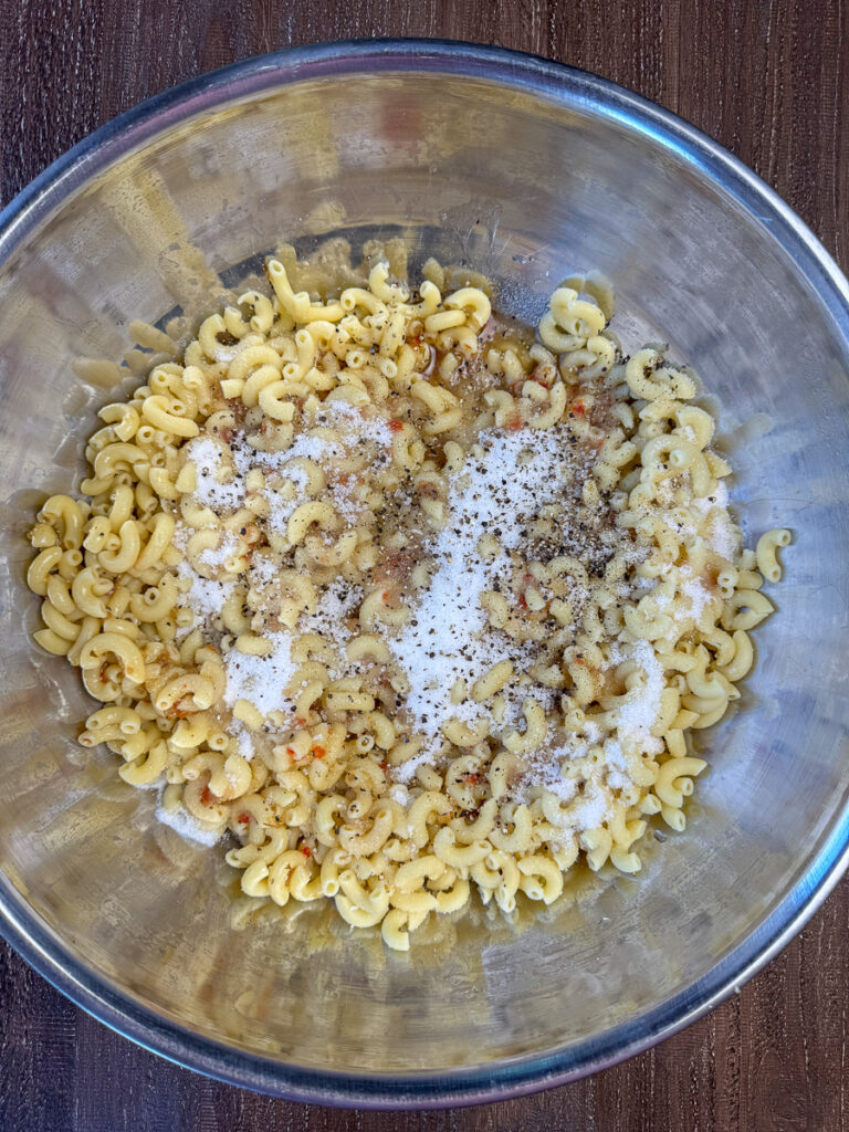 A bottle of Italian dressing poured over cooked elbow macaroni with sugar, salt and pepper sprinkled on top.
