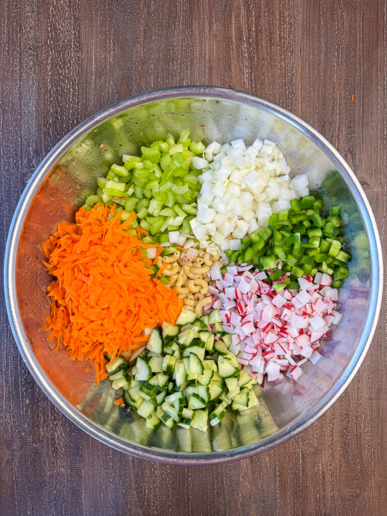 Shredded carrots, sliced cucumbers, chopped onion, green peppers, radishes and cucumbers on top of a bowl of Italian Macaroni Salad.