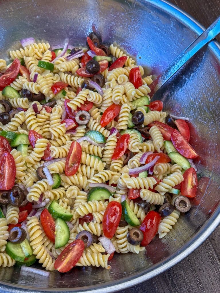 Silver spoon stirring together a mixing bowl full of rotini pasta, cherry tomatoes, cucumber, olives and red onion with a light, homemade Greek salad dressing.