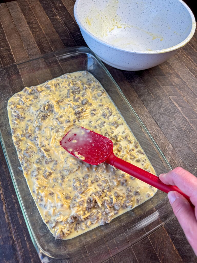 Spatula smoothing out the top of a souffle recipe in a glass dish on a wooden counter.