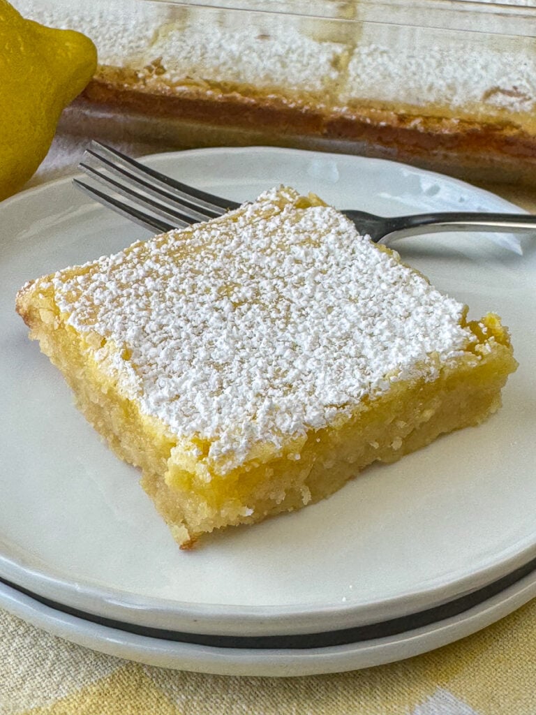 Cool, citrusy and refreshing lemon bar on a white plate with a fork ready to take a bite.