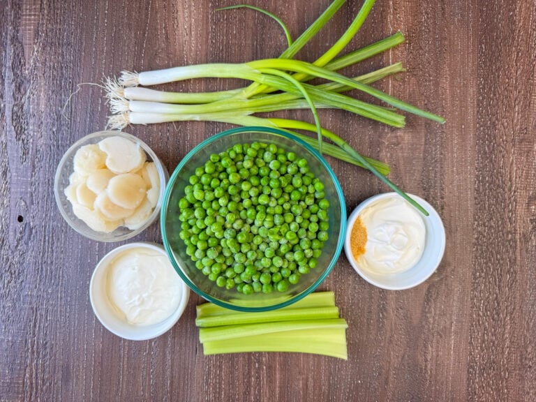 Ingredients to make old fashioned pea salad are green onions, sour cream, seasoned salt, peas, celery, mayonnaise and water chestnuts on a wooden counter top.
