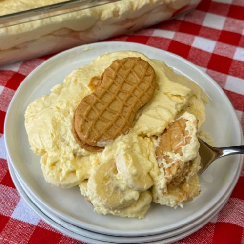 Close up of a spoon ready to lift a scoop of banana pudding with peanut butter cookies in an extra rich, cheesecake like pudding mixture off of a white plate.