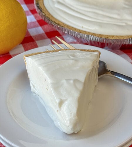 Creamy pie of frozen lemonade pie on a white plate with the full pie and a lemon in the background.