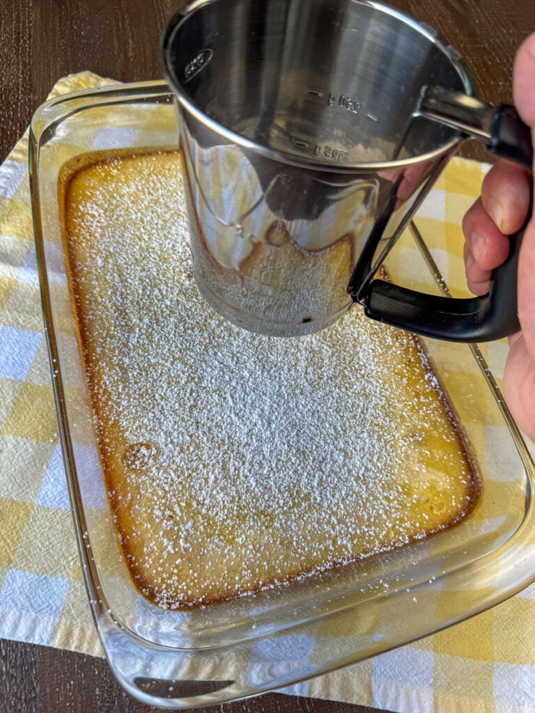 Powdered sugar being shaken over top of homemade lemon bars on a yellow and white plaid napkin.