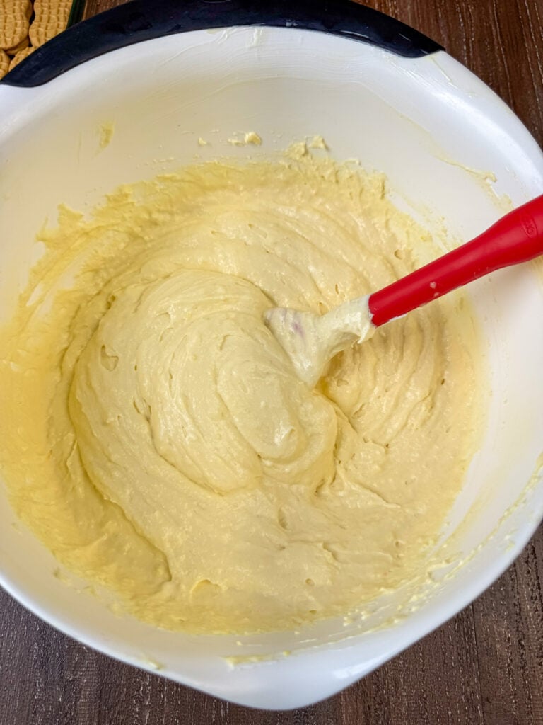 Spatula stirring instant vanilla pudding and milk into a rich mixture of cream cheese and sweetened condensed milk in a large white mixing bowl.