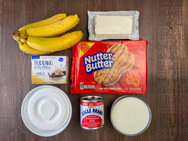 Ingredients to make Nutter Butter Banana Pudding are ripe bananas, instant vanilla pudding, cream cheese, Nutter Butter peanut butter cookies, milk, sweetened condensed milk and whipped topping on a wooden counter top.