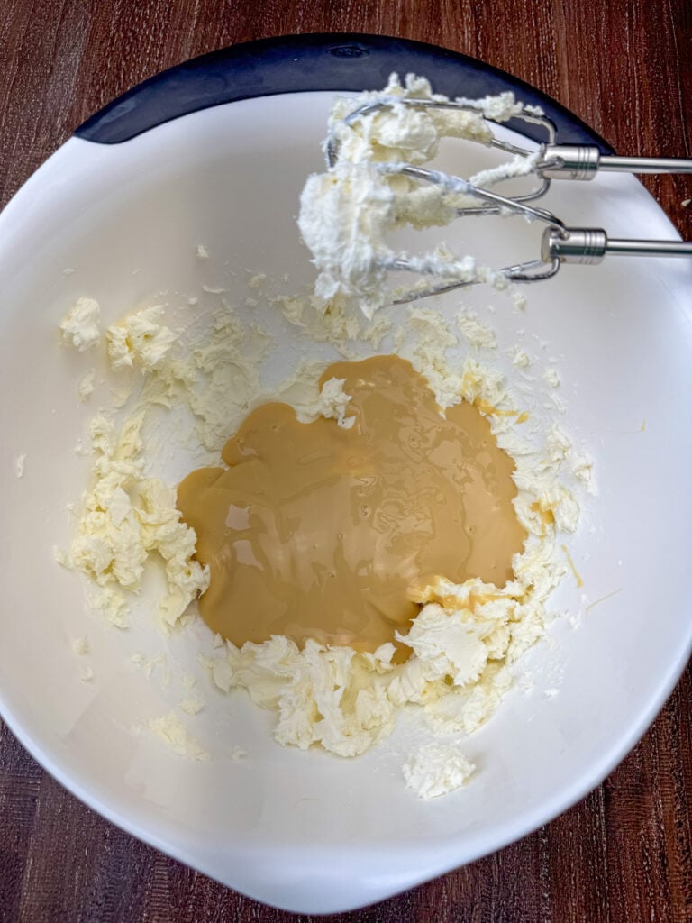 Beaten cream cheese in a mixing bowl with a can of sweetened condensed milk added to the bowl and beaters resting over the top edge.