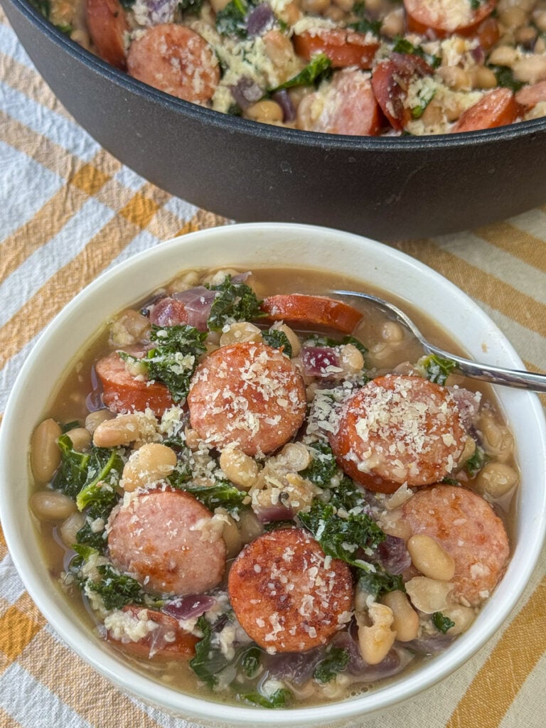 Bowl of sausage, kale and white bean recipe with a sprinkling of Parmesan and a spoon ready to scoop up a serving next to the skillet full.