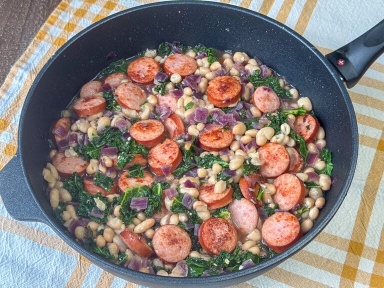 Side view of a twenty minute skillet meal with tender red onions nestled in a flavorful mixture of white beans, sausage and kale resting on a yellow and white napkin.