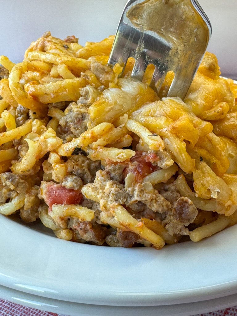 Close up of a fork in a serving of spaghetti in a creamy taco seasoned sauce filled with ground beef, onions and tomatoes then covered with melted cheese.