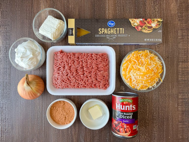 Ingredients to make taco spaghetti are sour cream, cream cheese, spaghetti, Mexican cheese, ground beef, diced tomatoes, butter, taco seasoning and an onion on a wooden counter top.