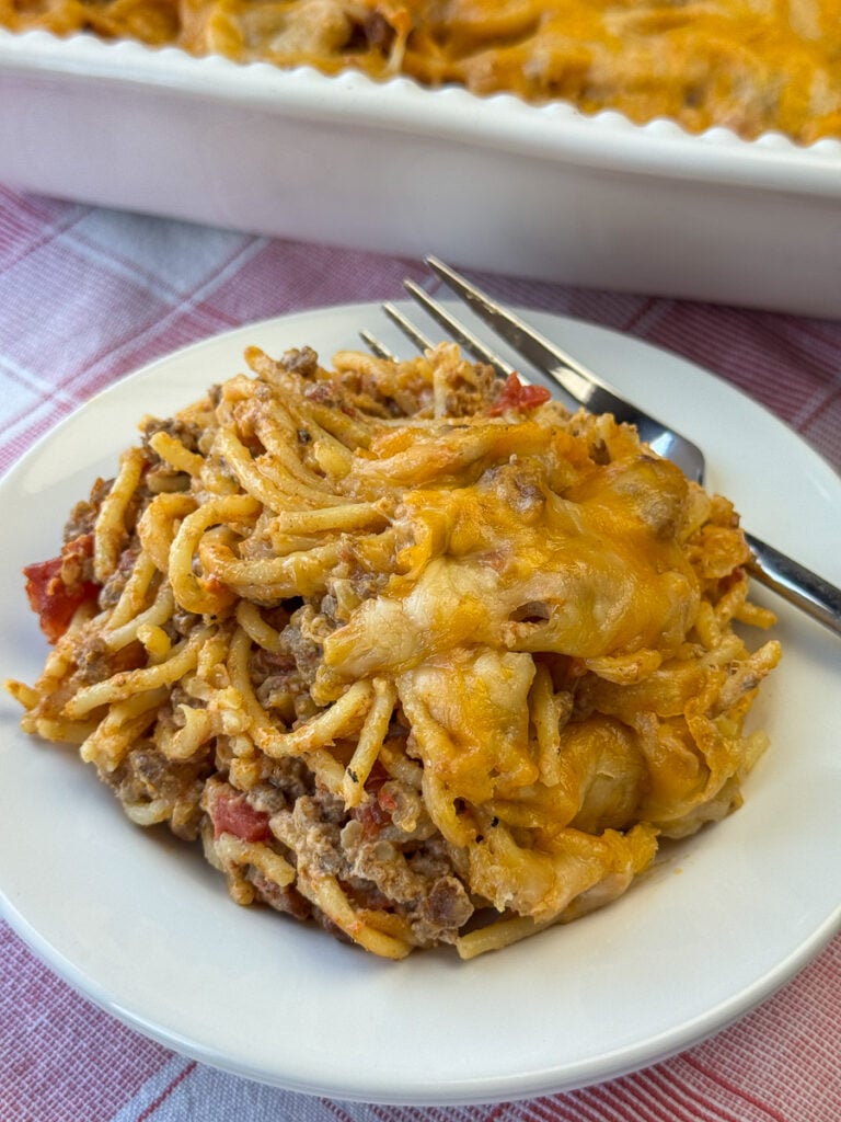 Overhead view of serving of taco spaghetti casserole on a plate with the full dish in the background. Most is covered with a Mexican blend cheese, but a portion is revealing generous amounts of ground beef, tomatoes and a few onions.