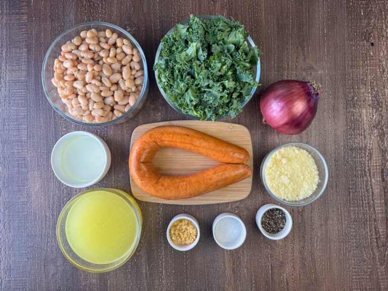 Ingredients to make a white bean and sausage skillet meal are navy beans, chopped kale, red onion, grated Parmesan cheese, Kielbasa sausage, vegetable oil, chicken broth, minced garlic, salt and pepper on a wooden counter top.
