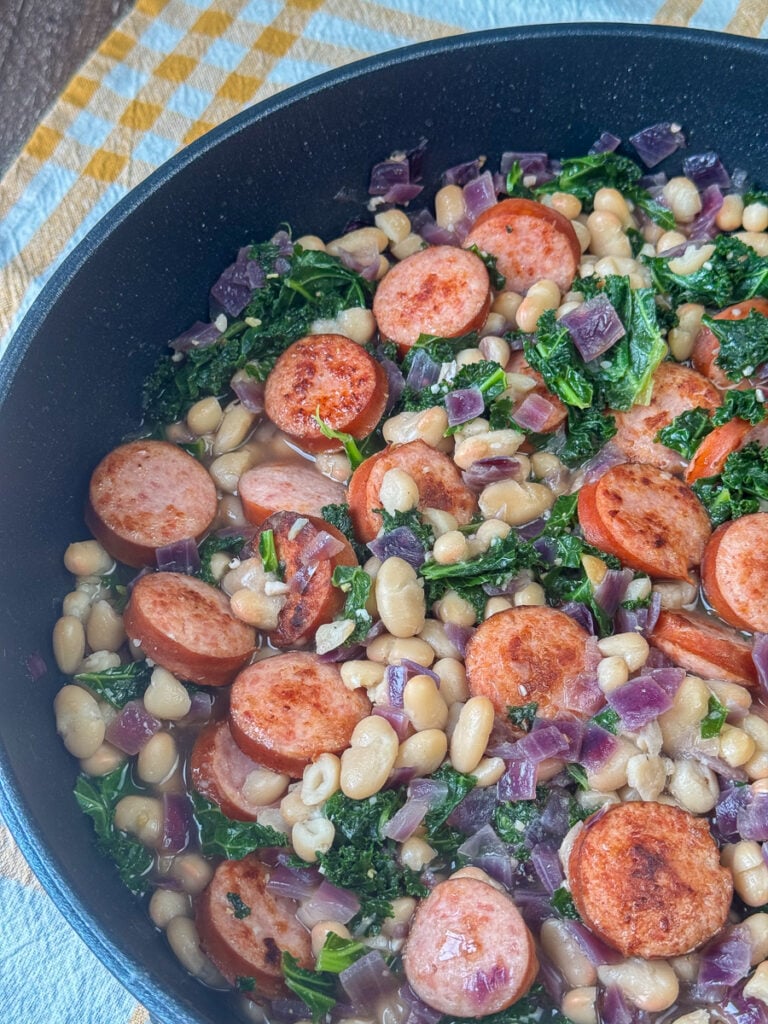 Close up view of tender navy beans cooked in a little broth with kale and sausage in a black pan.