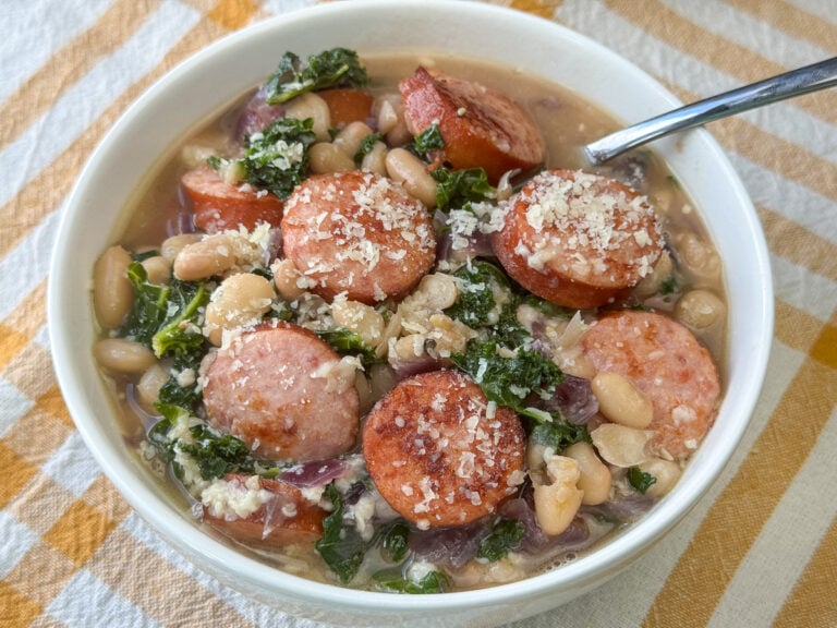 Close up bowl of browned slices of smoked sausage and white beans in a flavorful broth with bits of kale and red onion cooked in.