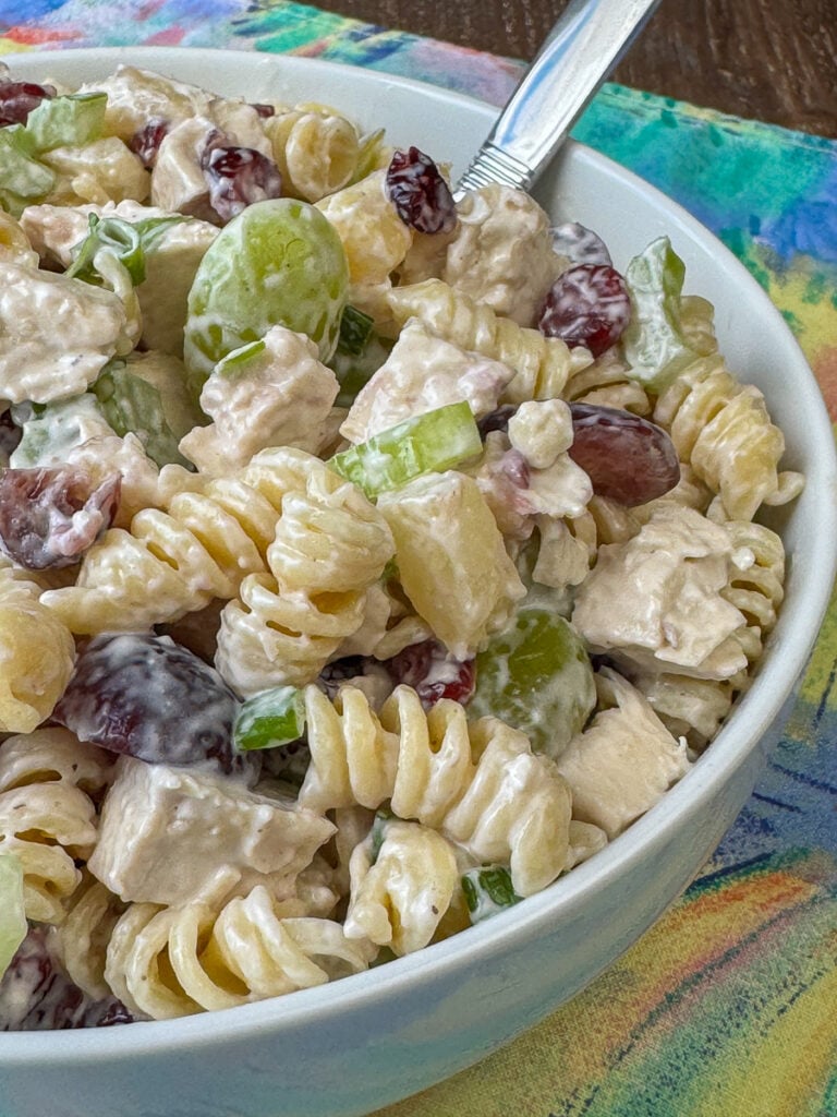 Close up of a refreshing chicken pasta salad with red grapes, pineapple, cashews, green onions, celery and green grapes with a silver serving spoon.