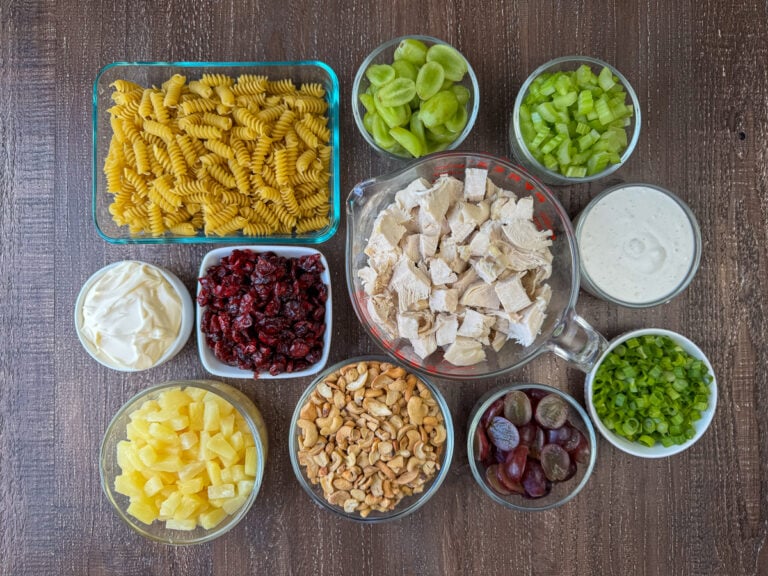 Ingredients to make cashew chicken pasta salad are rotini pasta, green grapes, sliced celery, ranch dressing, cubed chicken, dried cranberries, mayo, pineapple tidbits, cashews, red grapes and sliced green onions on a wooden counter top.
