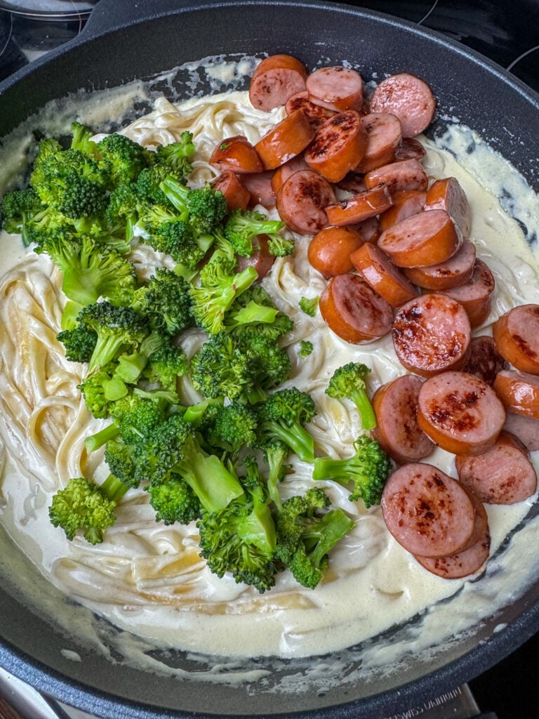 A pound of browned Kielbasa slices spread over fettuccine noodles in white sauce and cooked broccoli next to it.