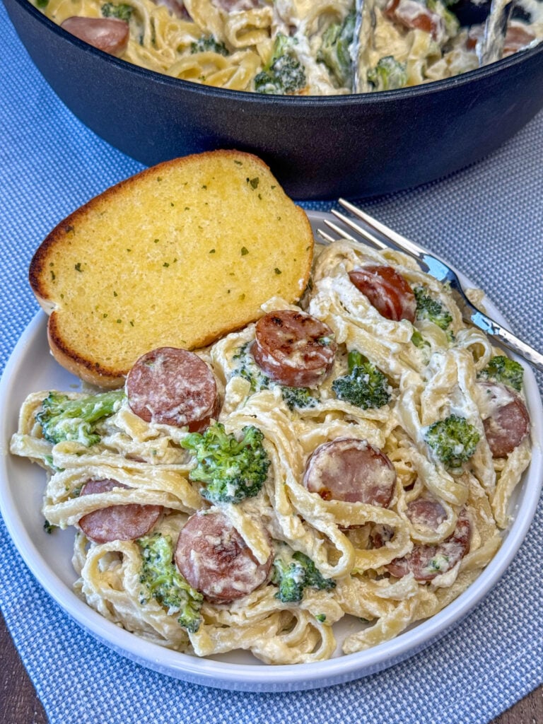 Plate of Fettuccine Alfredo with smoked sausage and broccoli and a slice of garlic bread next to the pot full of creamy pasta.