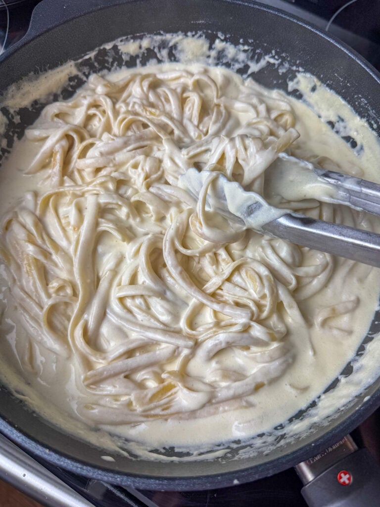Tongs stirring fettuccini noodles into a simple homemade Alfredo sauce in a skillet.