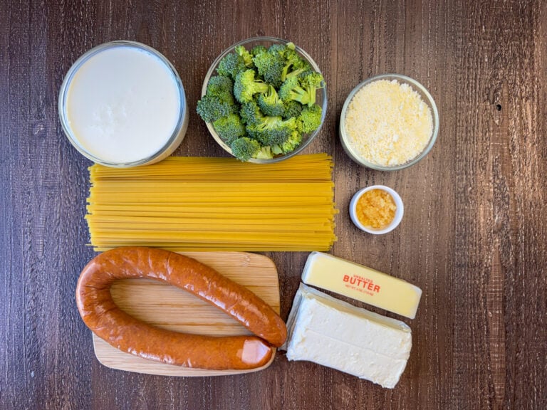 Ingredients to make Kielbasa and Pasta are heavy cream, fettuccine pasta, broccoli florets, grated parmesan, minced garlic, butter, cream cheese and Kielbasa on a wooden counter top.