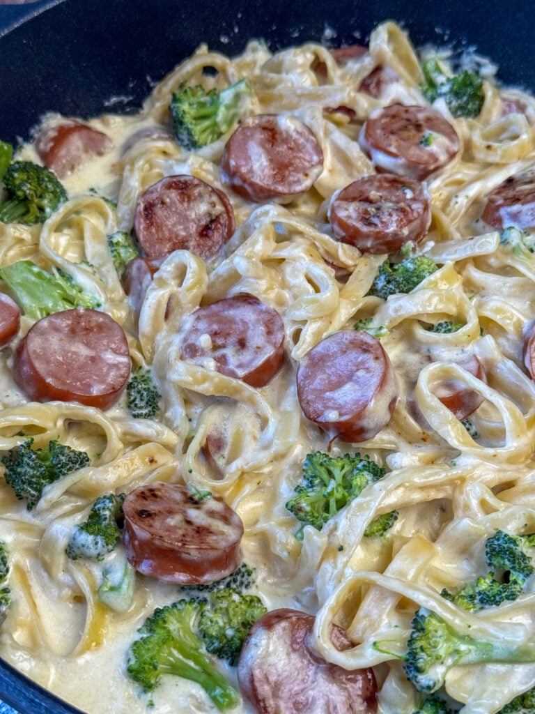 Skillet full of pasta generously coated in a creamy Alfredo sauce with broccoli and slices of Kielbasa sausage.