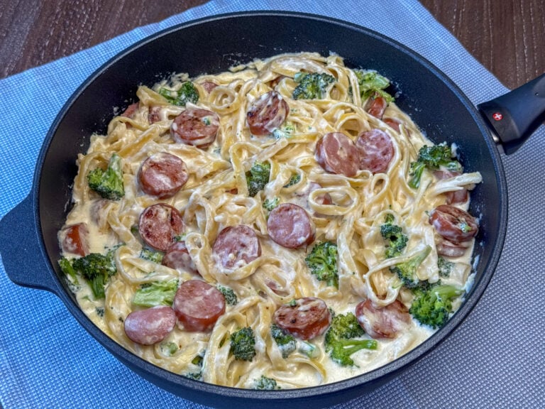 A warm mixture of pasta with plenty of Kielbasa stirred into a creamy butter and cheese sauce in a deep skillet on a blue napkin.